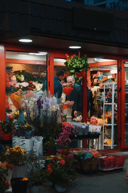 Exterior view of a flower shop near Romford Station, displaying a vibrant variety of fresh floral arrangements and bouquets inside the brightly lit storefront. Outside, numerous colorful potted plants
