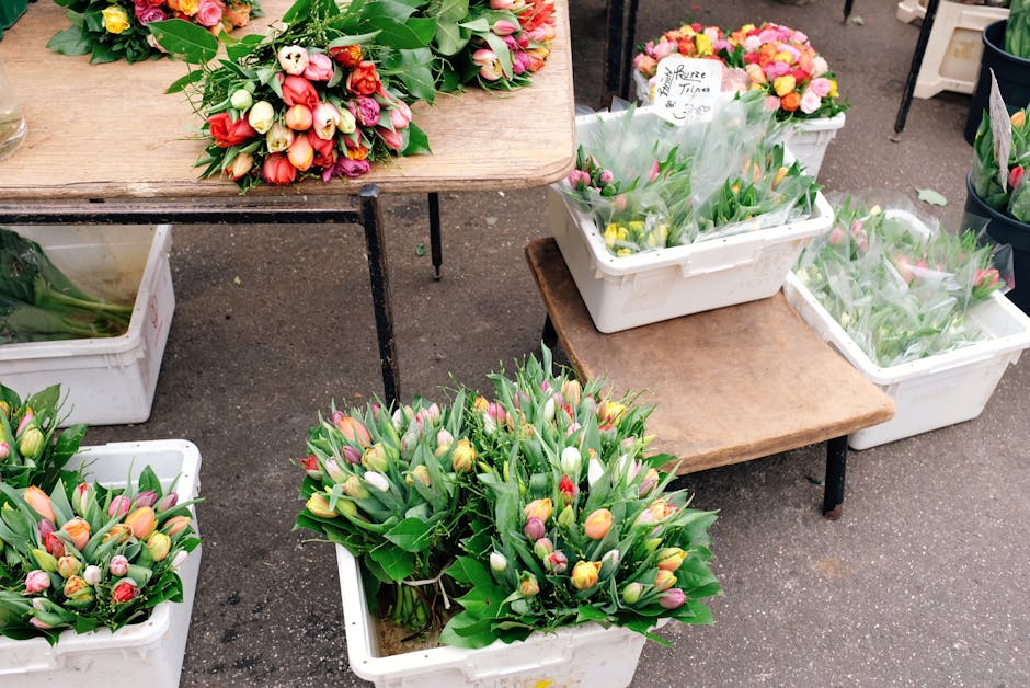A display of fresh tulip bouquets arranged in white plastic bins and placed on wooden tables at Romford Market. The bouquets feature tulips in a variety of colors, including pink, yellow, white, and r