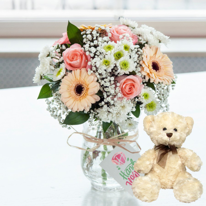 On a spotless white surface, a lovingly composed floral bouquet and a plush teddy bear form a gentle anniversary tableau that feels very at home in Romford's cosy living rooms. The clear glass jar vase at the centre cradles a rounded arrangement of flowers in soft, romantic tones. Pale pink roses unfurl their satin-like petals beside warm peach gerbera daisies, whose deep brown centres add depth and contrast. Between them, delicate white baby's breath spills like lace, while button-sized white chrysanthemums with lime-green centres bring a light, refreshing note, reminiscent of spring mornings in Romford's neighbouring parks. The stems are visible through the vase, neatly trimmed and fanning out in the water, underscoring the freshness of the arrangement. Around the neck of the vase, a slim length of natural twine is tied with an effortless bow, from which a small white tag hangs. The tag is decorated with a simple pink motif and the word "Handmade" in flowing green script, hinting that this bouquet has been created with a personal touch, as local florists in Romford often pride themselves on doing for special anniversaries. To the right of the vase, a cream teddy bear sits upright, positioned slightly at an angle toward the flowers. The bear's plush fur looks wonderfully soft and comforting, and its features are classic and understated: dark brown bead eyes, a tiny matching nose, and a neatly knotted brown satin ribbon at the neck that adds a hint of elegance. The entire scene is illuminated by soft, bright natural light from a blurred background, suggesting a daylight-filled room or a window looking out toward Romford's residential streets. Subtle reflections on the tabletop echo the pastel hues and the teddy's creamy coat, creating a warm, heartfelt image ideal for anniversary surprises, romantic gestures, or any occasion where tenderness and thoughtfulness are the message.